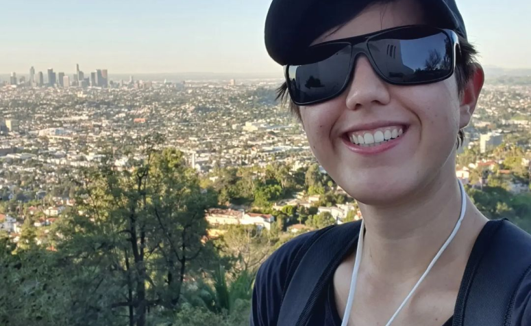 mulher com cabelos curtos e escuros, pele clara, usando óculos escuros, tirando selfie, ao fundo paisagem verde