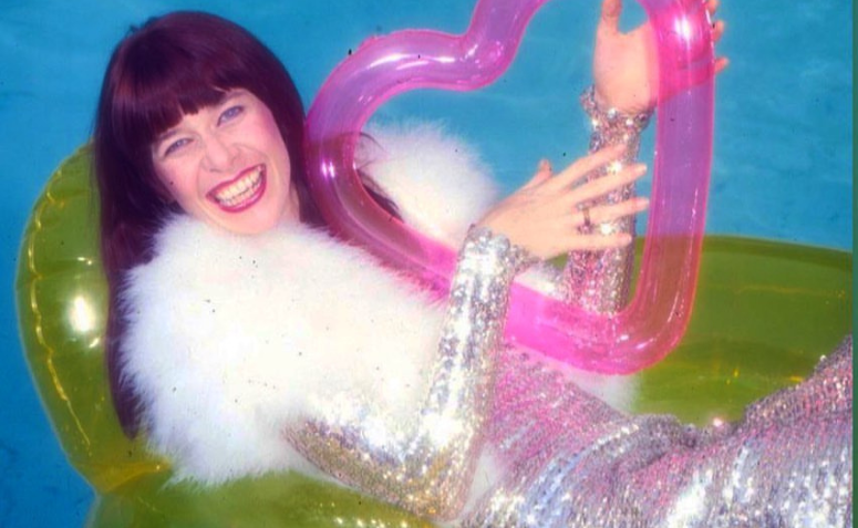 rita lee jovem sorrindo, com cabelo comprido e roupa prateada com ombreiras de pelo sintético branco. ela está em uma boia, em uma piscina, segurando uma boia pequena de coração como se fosse uma guitarra